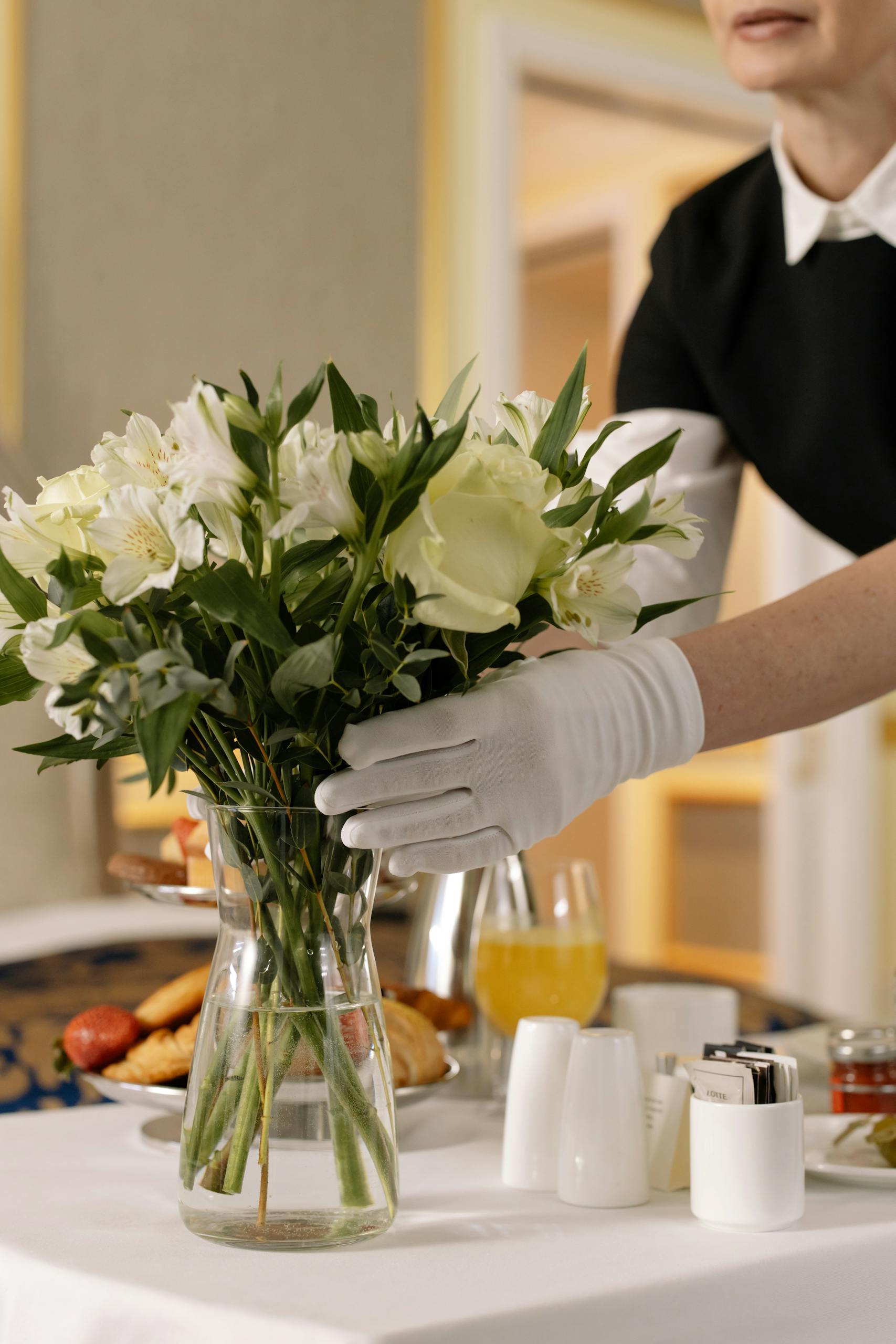 Woman arranging flowers in a hotel room with breakfast on a table, emphasizing luxury and convenience.